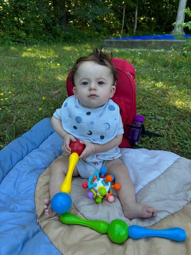 A baby sits up on a blanket in the outdoor space, holding a rattle, with more toys within reach.