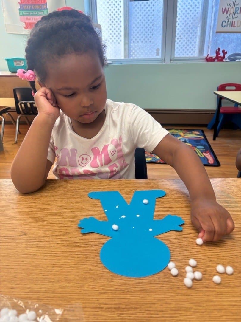 A preschooler presses cotton balls onto the paper snowman she is making at the craft table.