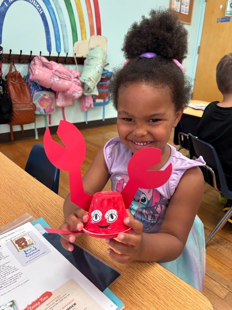 A child holds up the paper cup crab she made, smiling, with the coat hooks and painted rainbow behind her.