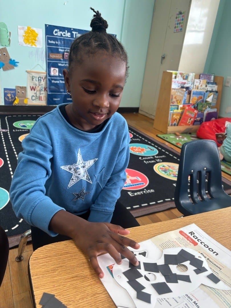 A preschooler places paper shapes on a raccoon mask from that week's lesson, beside the Circle Time board.