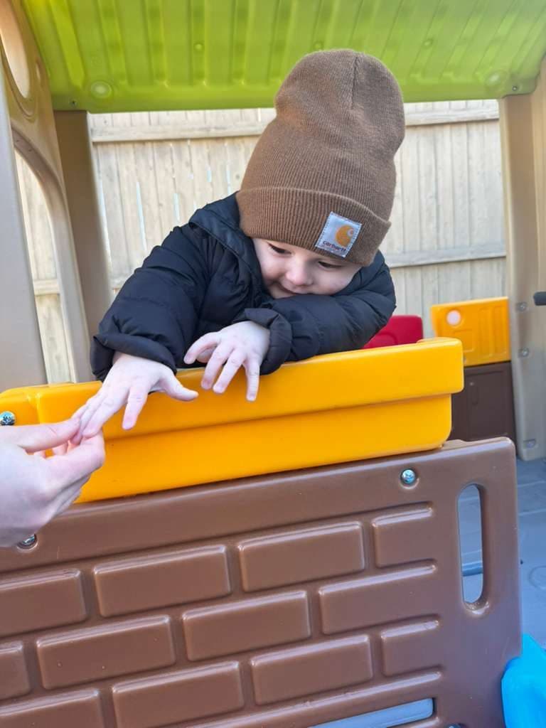 A toddler in a winter hat and coat leans over the wall of the outdoor playhouse to reach a teacher's hand.