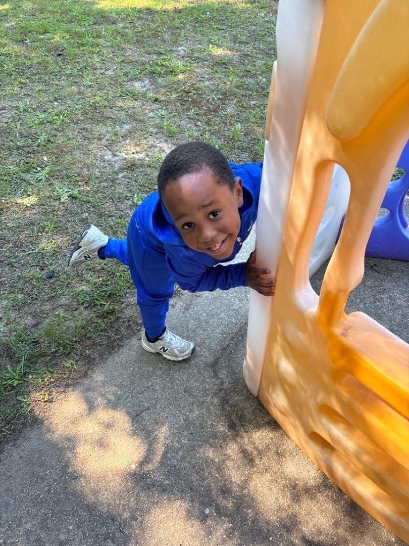 A child leans around the side of the playground slide, grinning at the camera.