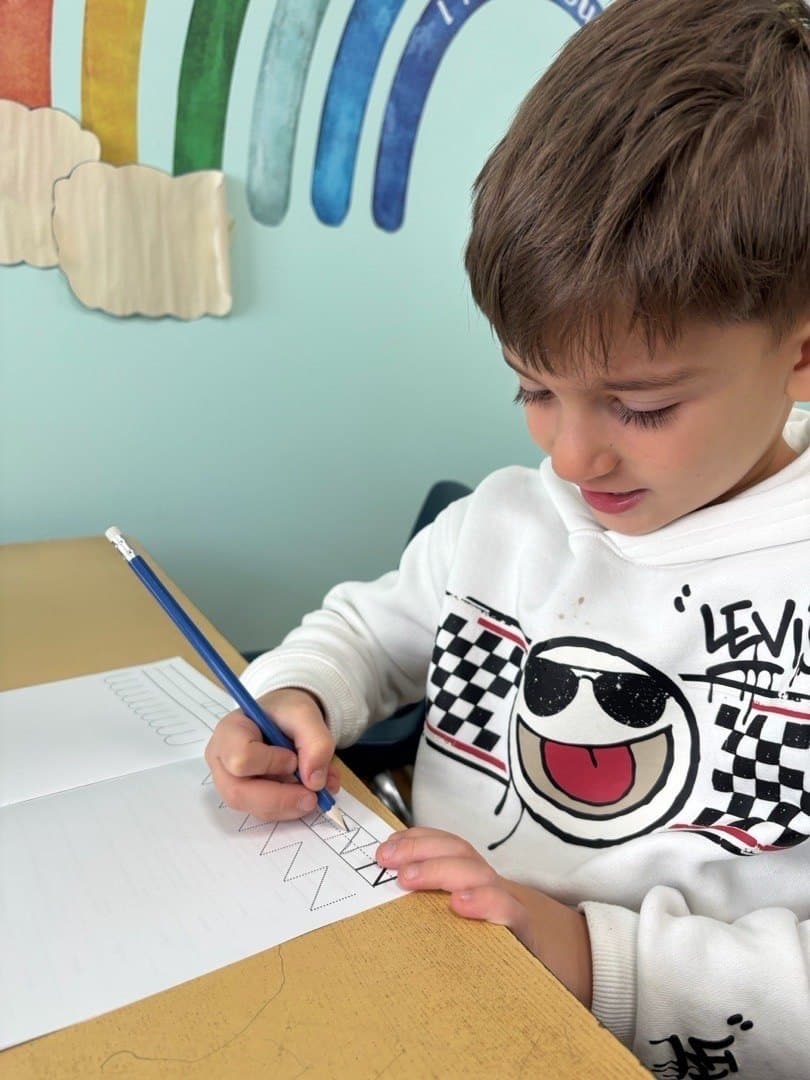 A pre-kindergarten child forms letters with a pencil on a tracing sheet, under the painted rainbow on the classroom wall.