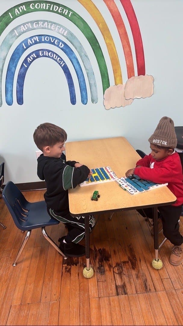 Two children work on letter and number boards at a table, below a painted rainbow reading I am confident, I am grateful, I am loved, I am enough.