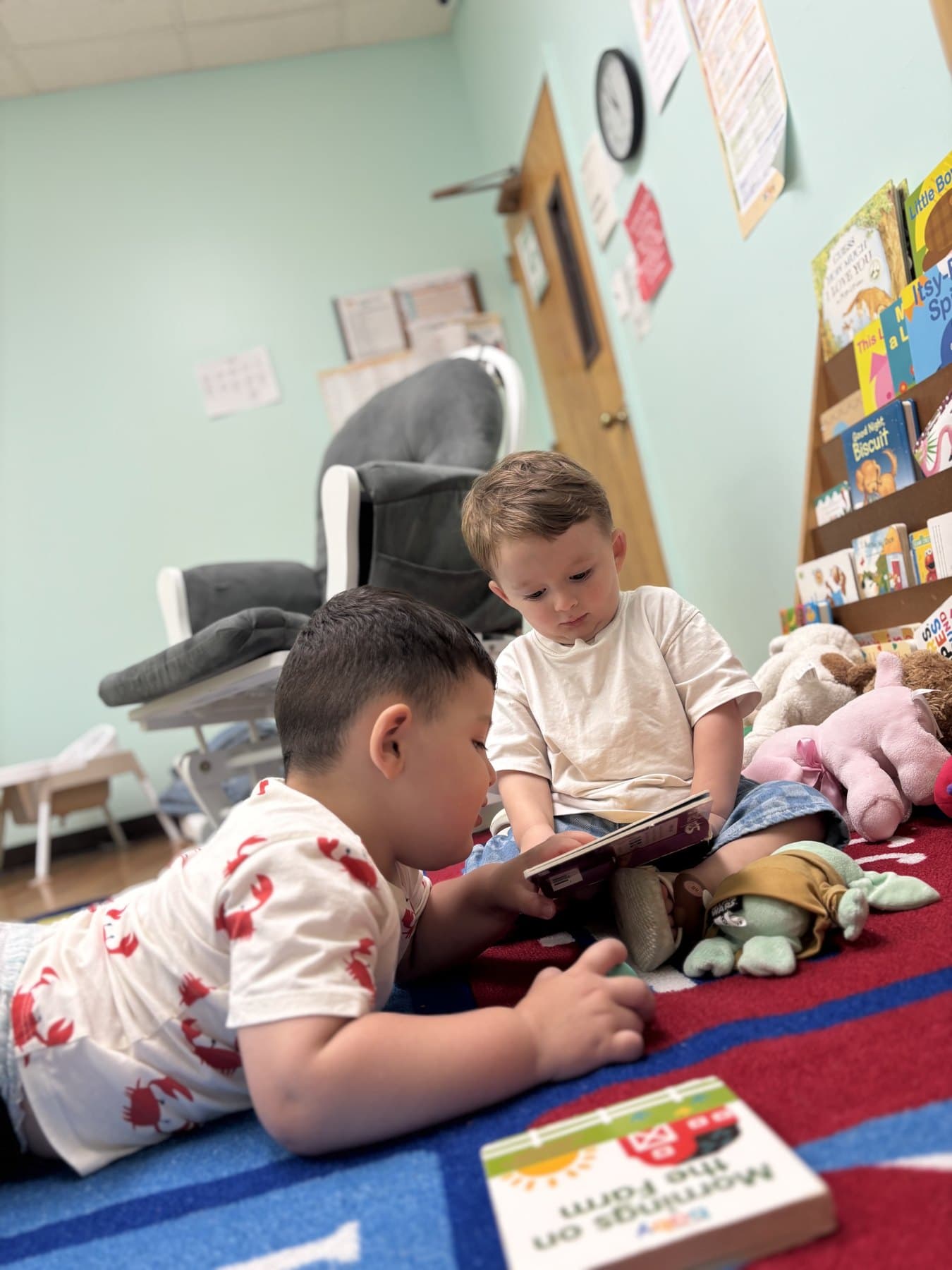 Two children share a board book on the rug in the reading corner, in front of the picture book rack.