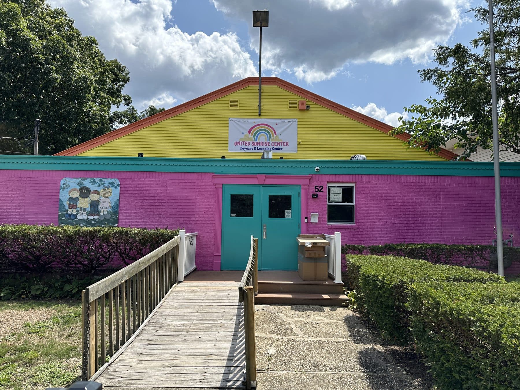 The front of the United Sunrise Center, pink brick under a yellow gable, with a mural of children beside the teal doors.