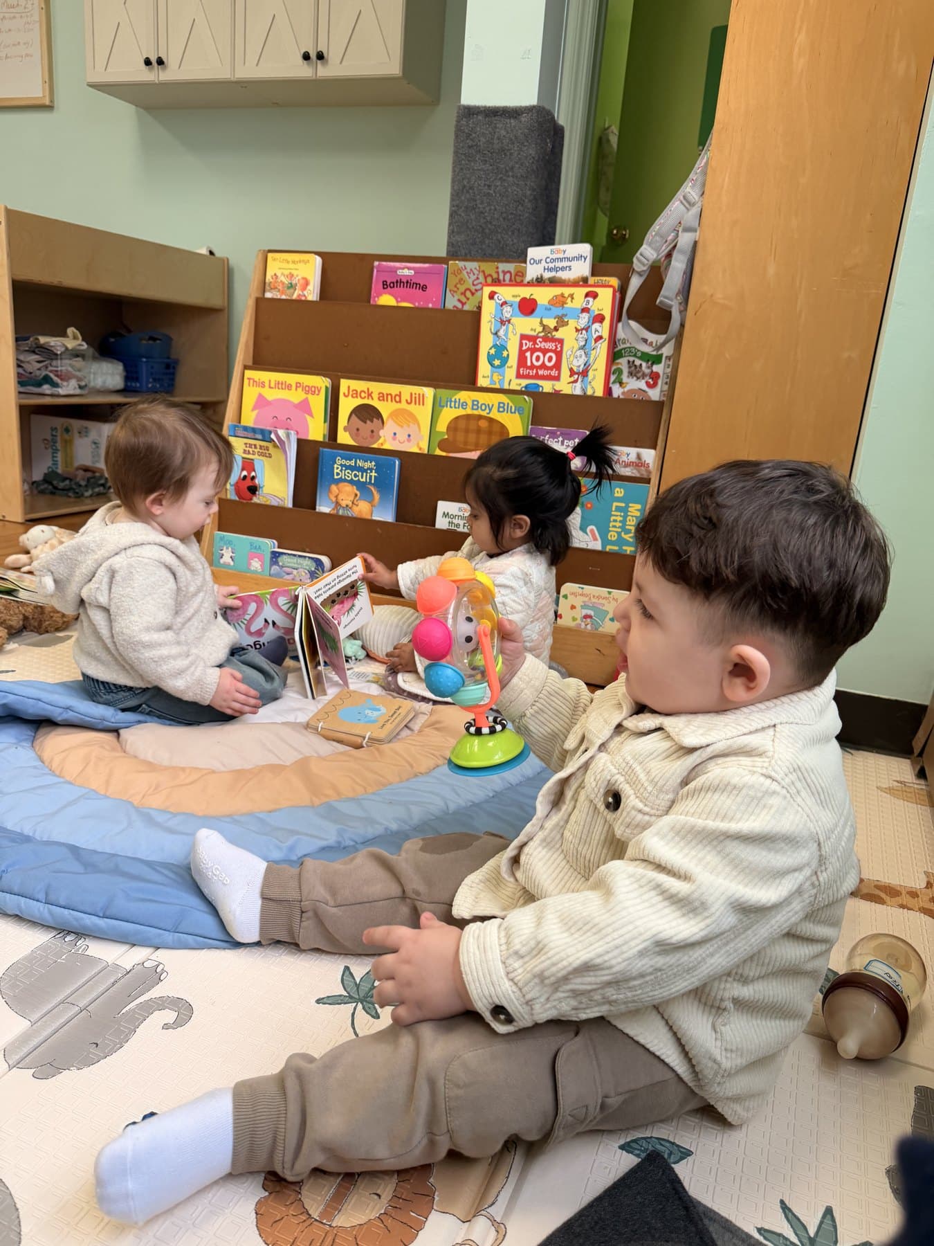 Three babies sit together on a play mat beside the board book rack in the infant room.