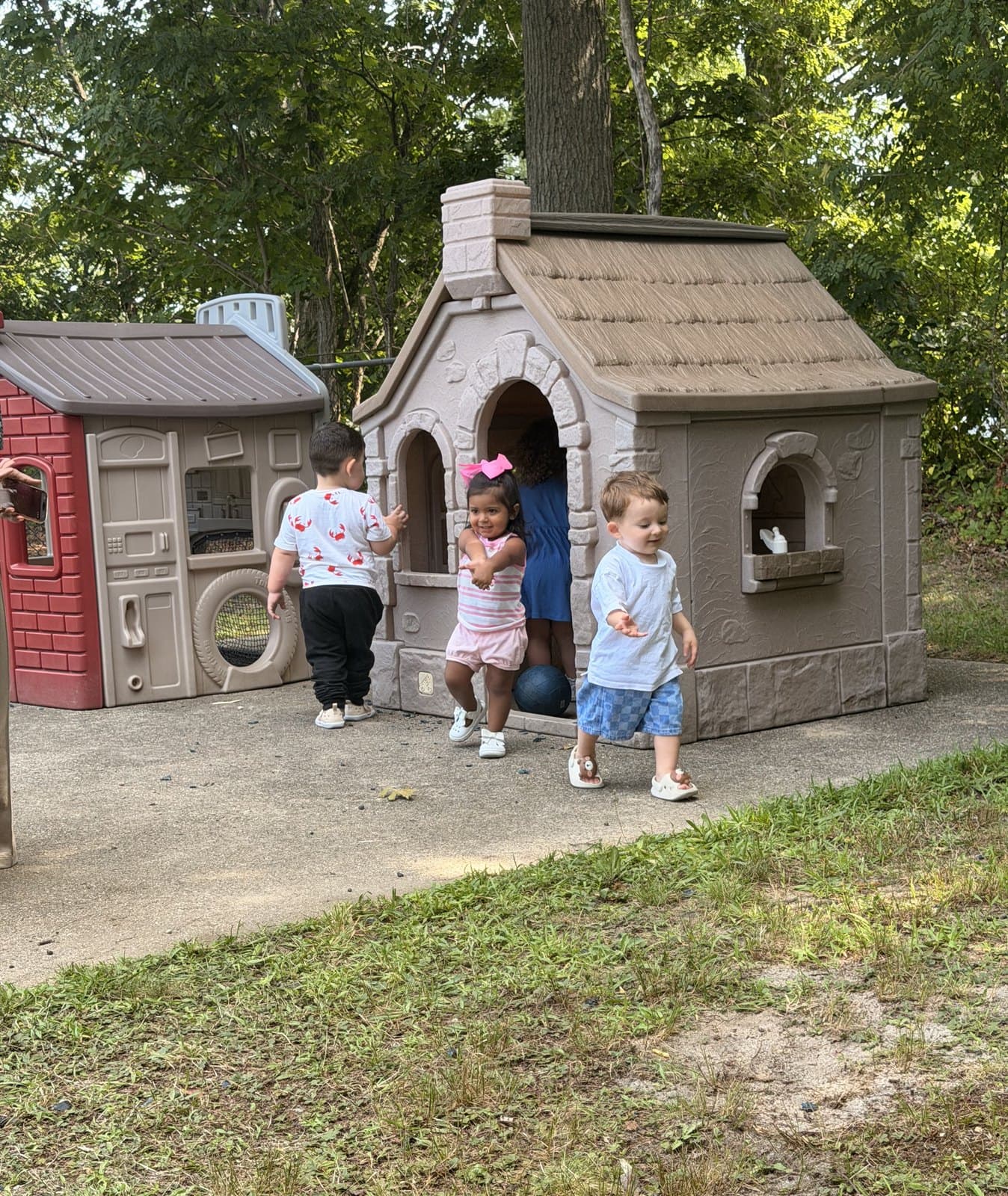 Four toddlers play around the two outdoor playhouses under the trees.