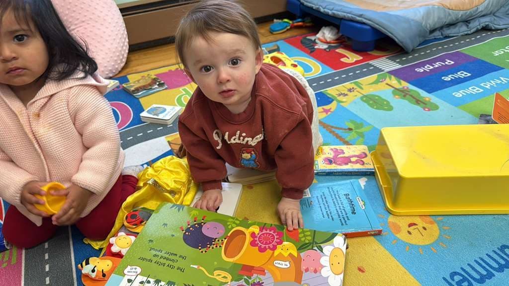 Two toddlers sit together on a colorful rug surrounded by open board books.